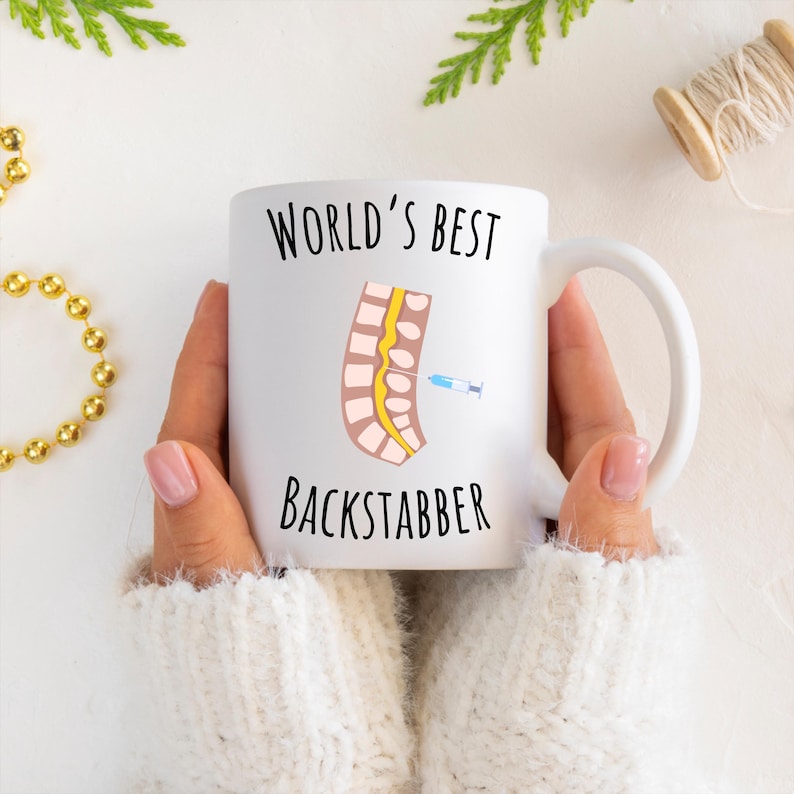 11 oz white glossy mug, Worlds Best Backstabber in black font, picture of blue and gray needle going into the spine, spine is tan color, spinal cord is yellow, depicting the spinal anesthesia process