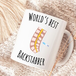 11 oz white glossy mug, Worlds Best Backstabber in black font, picture of blue and gray needle going into the spine, spine is tan color, spinal cord is yellow, depicting the spinal anesthesia process