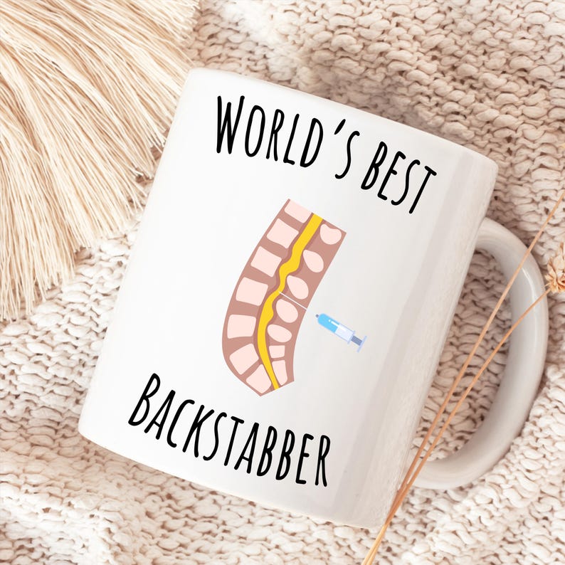 11 oz white glossy mug, Worlds Best Backstabber in black font, picture of blue and gray needle going into the spine, spine is tan color, spinal cord is yellow, depicting the spinal anesthesia process