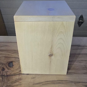 Op de afbeelding: Een vierkante houten doos van licht hout met een glad, onafgewerkt oppervlak. De doos is gemaakt van natuurlijk hout met zichtbare houtnerfpatronen.