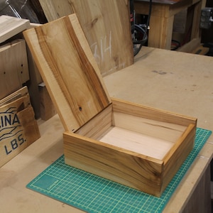 May include: A wooden box with a hinged lid, made of light brown wood with a natural wood grain pattern. The lid is open, revealing the empty interior of the box.