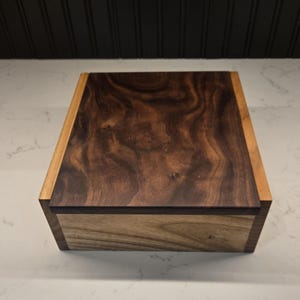 Könnte beinhalten: Eine Holzkiste mit einem dunkelbraunen Nussbaumdeckel und einem helleren Holzsockel. Der Deckel hat ein natürliches Holzmaserungsmuster.