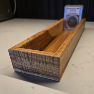 May include: A rectangular wooden tray with a visible natural grain pattern and mitered corners. Inside the tray, at the far end, sits a clear plastic rectangular case containing a circular object with a visible label and metallic details.