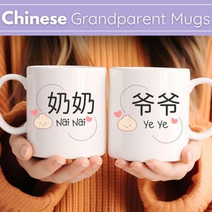 Peut inclure: Deux mugs en céramique blanche avec des caractères chinois noirs et des traductions anglaises. L'un dit "Nai Nai" avec une illustration de ravioli, l'autre dit "Ye Ye". Les deux mugs ont un cœur et un design en cercle pointillé. Les mugs sont tenus par une personne portant un pull orange.