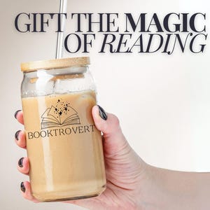 Puede incluir: Un tarro de vidrio con tapa de bambú y una pajita, lleno de un líquido marrón claro. El tarro tiene un logotipo en blanco y negro que dice "Booktrovert" y un diseño de un libro abierto con estrellas. El texto "Gift the Magic of Reading" está escrito en negro sobre un fondo blanco.