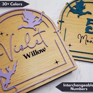 May include: Wooden sign with a purple dragon design and the name "Violet Willow" in black lettering. The sign has a light wood background and a purple border. The sign also has a small star design. The sign is for a baby's room.