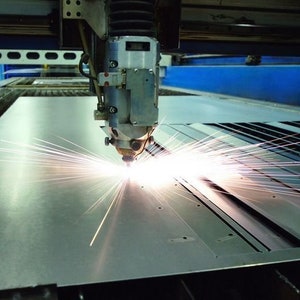 May include: A laser cutting machine in action, with bright sparks emanating from the cutting head as it cuts through a metal sheet. The machine is silver and grey, set against a blue and grey industrial background.