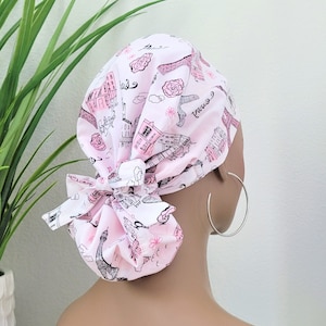 May include: A pink and white bouffant scrub cap featuring a Parisian theme. The cap is adorned with illustrations of the Eiffel Tower, buildings, roses, and the word "Paris". The cap is tied in a bow at the back.