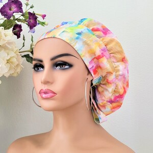 May include: A colorful surgical scrub hat with a pink, yellow, blue, and green abstract pattern. The hat is made of a soft, comfortable fabric and has a tie at the back.