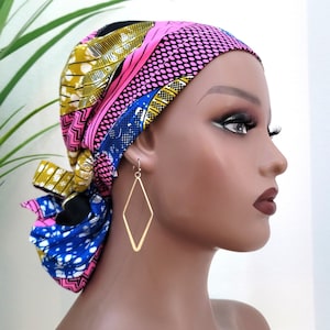 May include: A mannequin head is adorned with a vibrant, patterned head wrap in pink, blue, yellow, and black. Gold-colored diamond-shaped earrings dangle from the ear. The head wrap features a mix of geometric and floral designs.