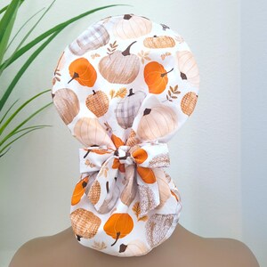 Pode incluir: Uma touca cirúrgica branca com um padrão repetido de abóboras em tons de laranja, bege e creme. A touca tem um laço nas costas, também com estampa de abóboras e folhas. Perfeita para a estação do outono.
