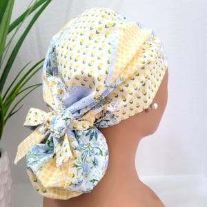May include: A colorful, patterned scrub cap featuring a mix of floral, gingham, and solid fabric panels. The cap is predominantly yellow, blue, and white, with a bow at the back. The design includes small yellow flowers and blue floral patterns.
