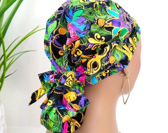 May include: A black scrub hat with a colorful Mardi Gras themed print featuring gold masks, purple, green, and gold feathers, and gold fleur-de-lis. The hat is tied in the back.