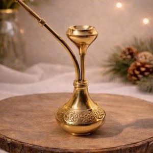 May include: A gold-colored hookah with an ornate, engraved base and a curved stem. The hookah sits on a wooden surface, with a blurred background featuring greenery and soft lighting. The design is reminiscent of Middle Eastern decor.