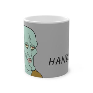 Puede incluir: Una taza de cerámica gris con una imagen de dibujos animados de un personaje de piel verde con ojos rojos y una camisa marrón. El texto "HANDS" está impreso en negro en la taza.