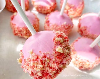 May include: Pink cake pops with a strawberry crumble coating, presented on white sticks. The pops are arranged in a white container, showcasing a dessert treat. The image features a close-up view of the treats.
