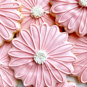 Puede incluir: Galletas rosas con forma de margarita con detalles de glaseado blanco y un centro de chispas blancas. Las galletas están decoradas con un glaseado rosa suave y líneas blancas para crear los pétalos.