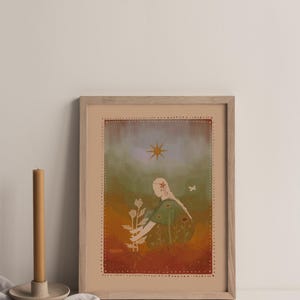 May include: Framed art print featuring an illustration of a person tending to a plant. The artwork has a warm color palette with a figure in a green dress, a star, and a butterfly. The print is in a wooden frame.