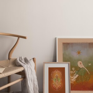 May include: Two framed art prints with illustrations, one featuring a woman with flowers and the other a figure with a halo. A wooden chair with a woven seat and a draped, light-colored fabric are in the foreground.