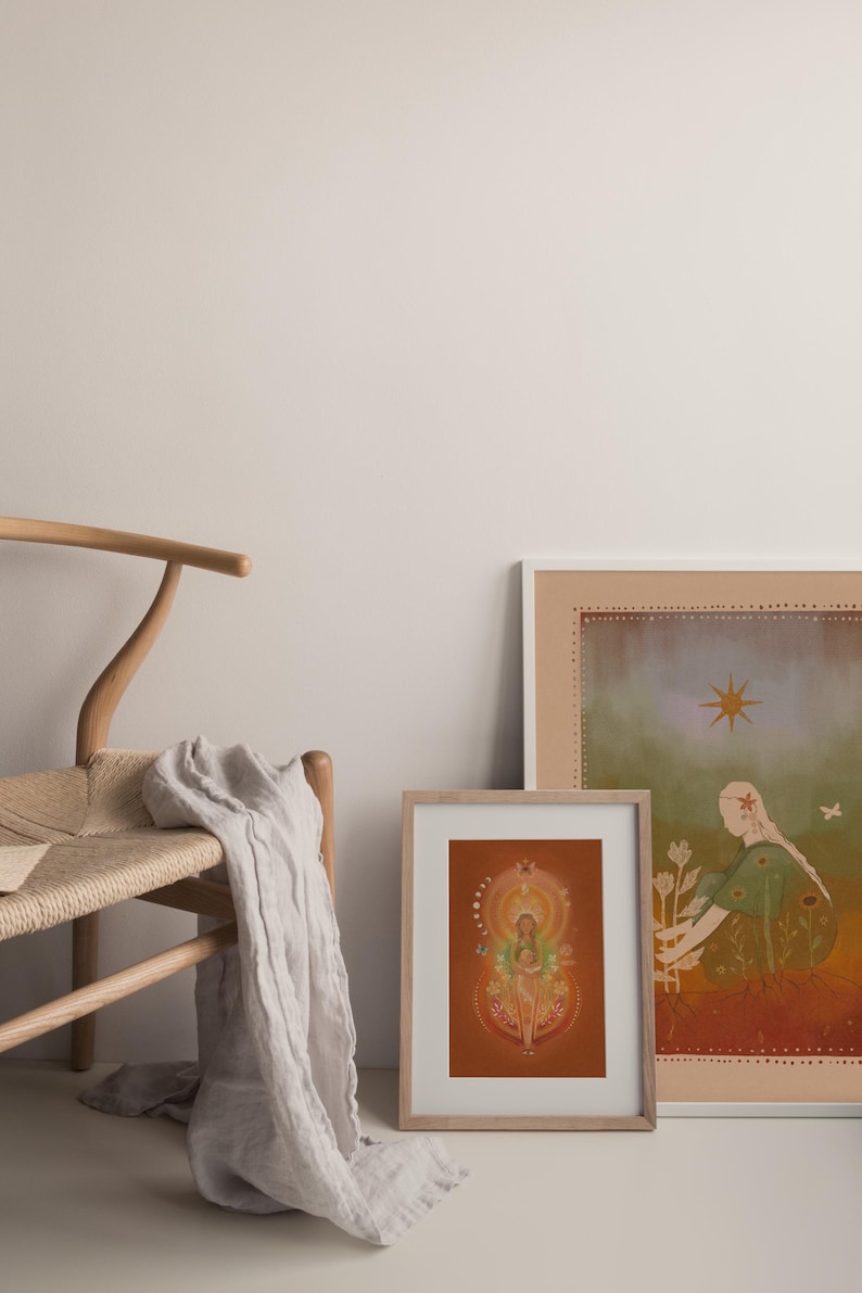 May include: Two framed art prints with illustrations, one featuring a woman with flowers and the other a figure with a halo. A wooden chair with a woven seat and a draped, light-colored fabric are in the foreground.