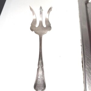 May include: A silver-colored serving fork with a trident-shaped head. The fork has three tines with curved edges and a long, slender handle. The handle has a decorative design near the base.
