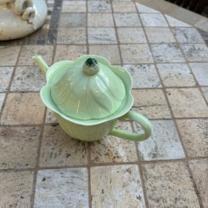 May include: A light green ceramic teapot with a flower-shaped lid and a green and brown flower detail on top. The teapot is sitting on a tiled surface.