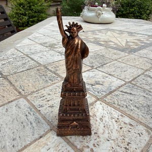 May include: A bronze-colored statue of the Statue of Liberty featuring a robed figure holding a torch aloft and a tablet in the other hand, standing atop a square, tiered pedestal base with detailed architectural carvings on all sides.