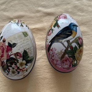Könnte beinhalten: Zwei dekorative Keramikeier mit Blumen- und Vogelmotiven. Ein Ei zeigt einen blauen Vogel, der auf einem Zweig mit rosa und gelben Rosen sitzt. Das andere Ei hat ein Vintage-Postkartenmotiv mit rosa Rosen und grünen Blättern.