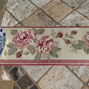 May include: Floral wallpaper border featuring large pink and red roses with green leaves and buds on a cream background. The border has a thin red stripe along the top and bottom edges. Decorative home decor.