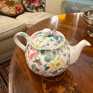 Könnte beinhalten: Eine weiße Teekanne mit einem Blumenmuster in Rosa-, Gelb- und Blautönen. Die Teekanne hat einen Deckel und einen Ausguss. Die Teekanne steht auf einem Holztisch.