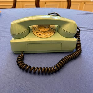 May include: A vintage, mint-green rotary dial telephone with a black, coiled cord. The phone has a classic design with a circular dial and a rectangular base. The dial features the numbers and the letters GTE.