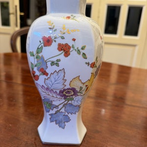Puede incluir: Un jarrón de porcelana blanca con un diseño floral. El jarrón tiene una forma geométrica y presenta flores rojas, amarillas, azules y moradas con hojas verdes. Las flores están delineadas en oro.