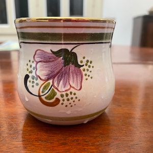 Könnte beinhalten: Eine weiße Keramiktasse mit goldfarbenem Rand und einem floralen Design. Das Design zeigt eine violette Blume mit grünen Blättern und braunen Akzenten.