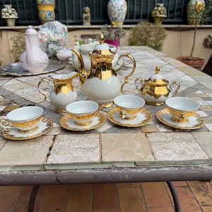 Può includere: Un servizio da tè bianco e oro con teiera, lattiera, zuccheriera e quattro tazze e piattini. Il set ha finiture e dettagli dorati. Il servizio da tè è esposto su un tavolo piastrellato.