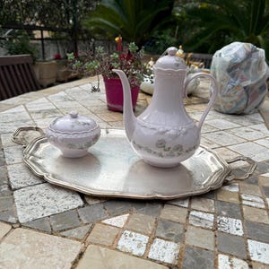 May include: A light pink porcelain tea set with floral detailing, comprising a teapot, sugar bowl, and a silver serving tray. The set is presented on a stone tile table, demonstrating a traditional aesthetic.