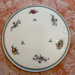May include: A round, white ceramic serving platter with a gold rim and a light blue border. The border features a repeating pattern. The platter is decorated with clusters of colorful flowers and fruits.