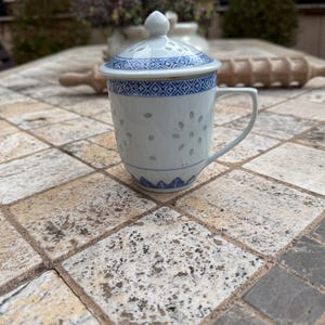 May include: A white ceramic teacup with a blue and white pattern and a lid. The teacup has a handle and a small hole in the lid. The teacup is sitting on a tiled surface.