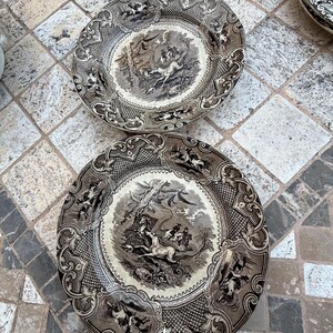 Può includere: Due piatti decorativi antichi con un design a scena di caccia in bianco e nero. I piatti hanno un bordo dettagliato con volute e ulteriori scene di caccia. I piatti sono rotondi e in ceramica.