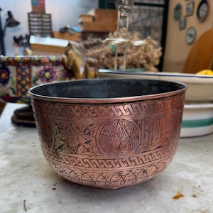 Puede incluir: Un cuenco de color cobre con intrincados patrones grabados. El cuenco tiene un interior oscuro y una forma ancha y redondeada. Los diseños grabados incluyen motivos geométricos y florales, añadiendo un elemento decorativo al recipiente.