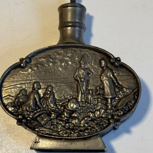 May include: A silver pewter bottle with a detailed embossed scene of people in a field. The bottle has a round body and a small, pointed stopper.