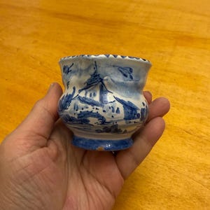 以下が含まれることがあります： 青と白のフラワーデザインが施された、小さな白い陶器のカップ。カップは丸みを帯びたボディと広がった縁を持っています。