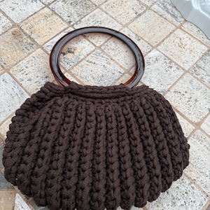 May include: A brown, handmade crochet handbag with a rounded shape. The bag features a dark brown, circular handle and a textured, ribbed design. The bag is sitting on a tiled surface.