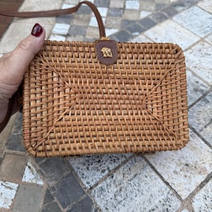 May include: A rectangular woven rattan handbag with a brown leather strap and a small leather accent featuring a decorative emblem. The bag is held by a person with dark red nail polish. The bag's dimensions are approximately 8 inches wide and 6 inches tall.
