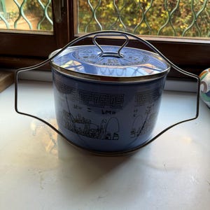 May include: A blue enamelware pot with a black and white design of people and a cart. The pot has a metal handle and a metal lid. The pot is sitting on a white surface.