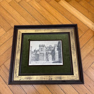 Può includere: Una stampa in bianco e nero incorniciata di una scena di città antica con un grande arco al centro. La cornice è di colore marrone scuro con una bordura dorata e un passepartout in velluto verde.