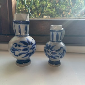 May include: Two blue and white ceramic pitchers with a floral design. The larger pitcher is taller and has a wider base. The smaller pitcher is shorter and has a narrower base.