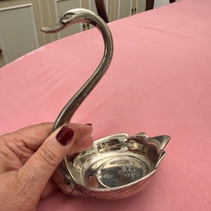 May include: A silver swan-shaped dish with a curved neck and a detailed head. The dish has a small, shallow bowl for holding items. The swan is held against a pink surface.