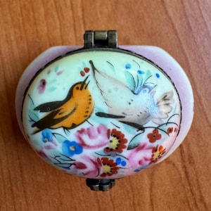 May include: A small, oval-shaped trinket box with a hinged lid. The box is painted with a delicate floral design and two birds, one yellow and one white, on a light blue background.
