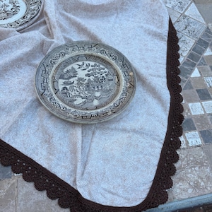 以下が含まれることがあります： 茶色の縁取りが施されたベージュ色のテーブルクロスと、2つのアンティークプレート。プレートには、伝統的な青と白のウィローパターンが描かれています。テーブルクロスは繊細な花柄です。