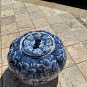 May include: A blue and white ceramic teapot with a lid. The teapot has a bulbous shape and is decorated with floral patterns. The lid features a small knob and a similar design. The teapot is sitting on a stone surface.